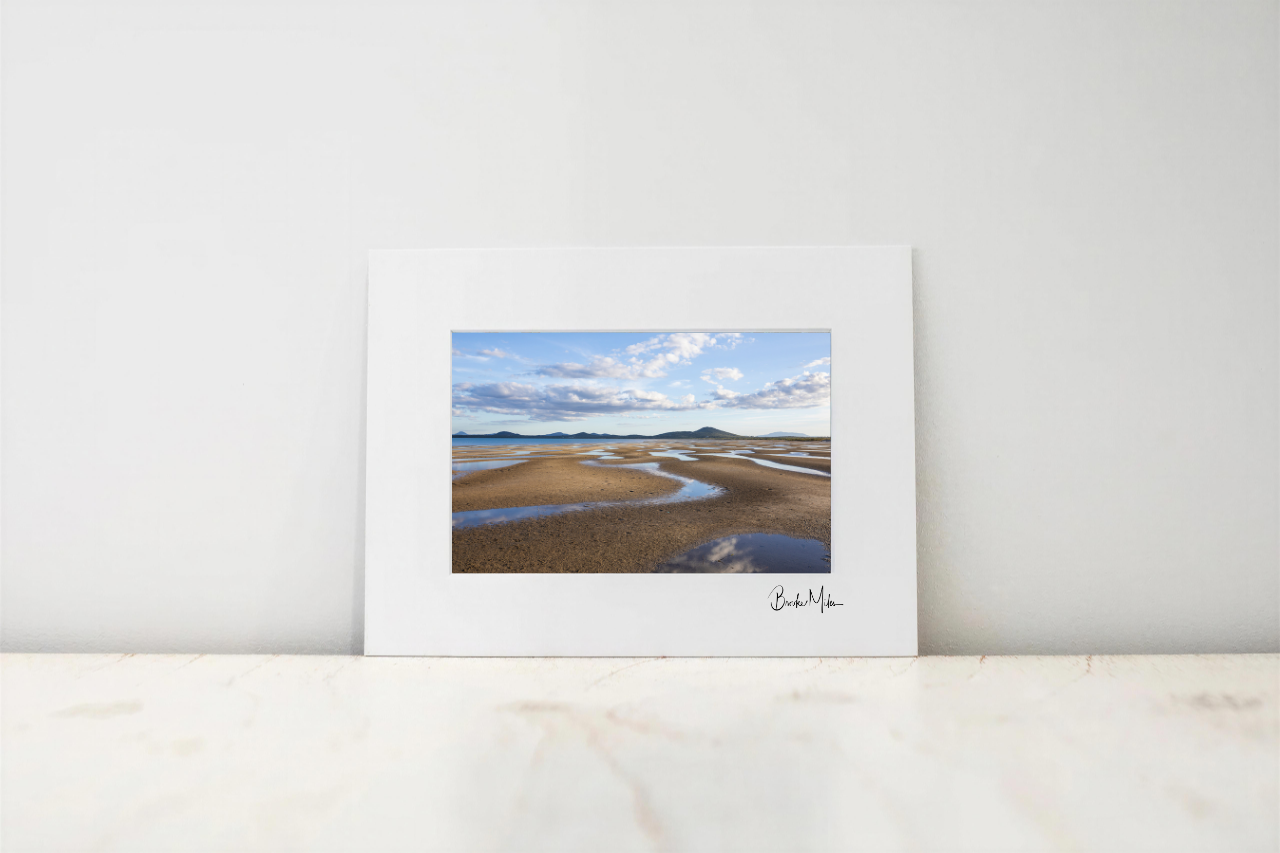A framed landscape art print featuring a tidal flat or beach scene with blue sky and clouds, displayed on a light marble surface against a white wall.