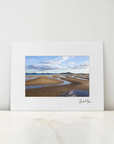 A framed landscape art print featuring a tidal flat or beach scene with blue sky and clouds, displayed on a light marble surface against a white wall.
