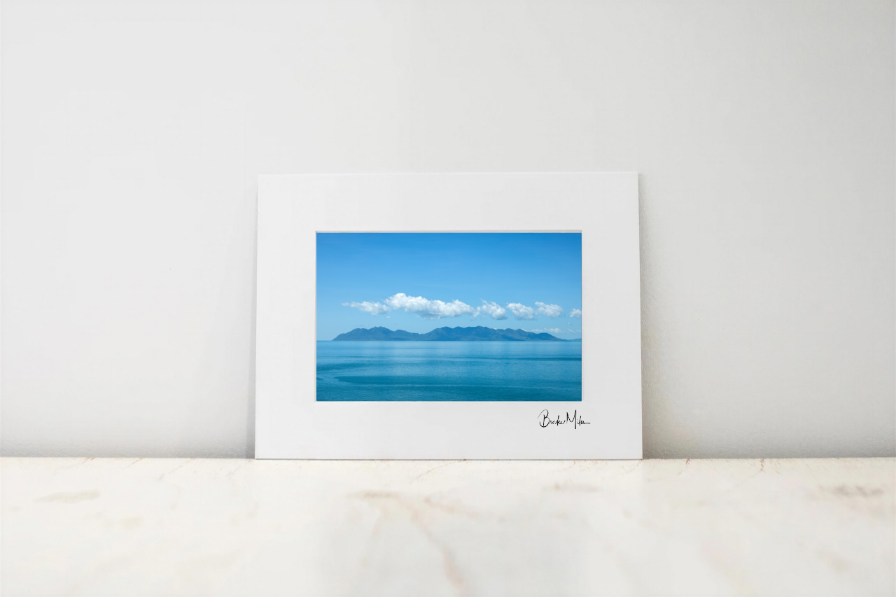 A framed art print featuring a scenic photograph of a blue sea and distant mountains under a clear sky, displayed on a light marble surface against a white wall.