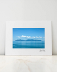 A framed art print featuring a scenic photograph of a blue sea and distant mountains under a clear sky, displayed on a light marble surface against a white wall.