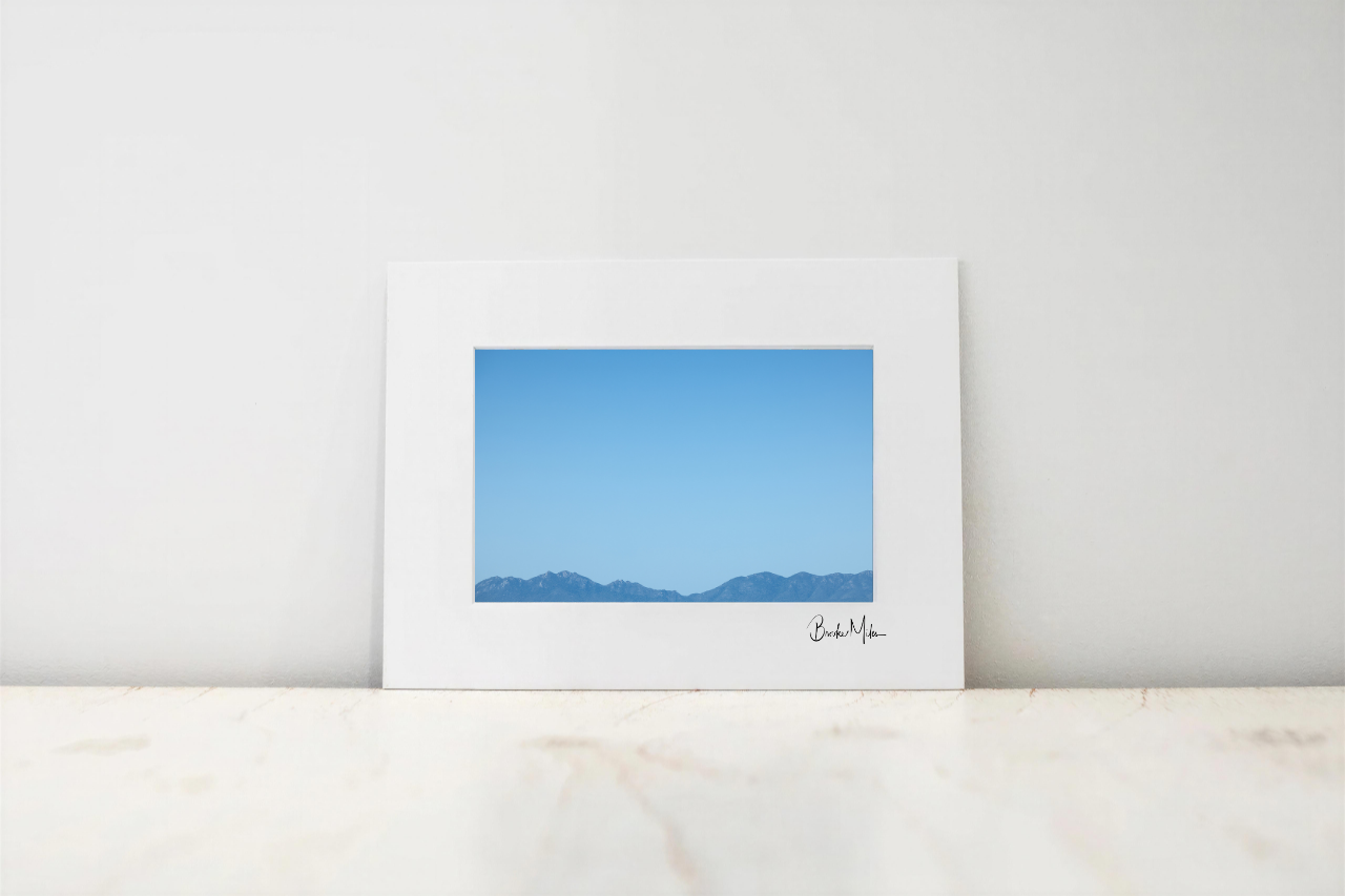 A framed minimalist landscape art print featuring a blue sky and distant mountain range, displayed on a light marble surface against a neutral white wall.