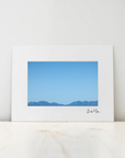 A framed minimalist landscape art print featuring a blue sky and distant mountain range, displayed on a light marble surface against a neutral white wall.