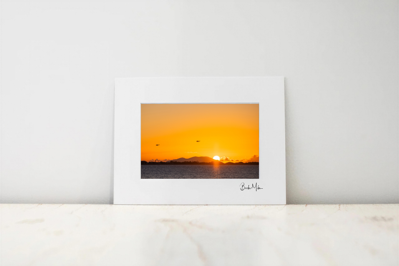 A framed art print featuring a vibrant orange and yellow sunset over water, displayed on a light marble surface against a neutral white wall.
