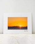 A framed art print featuring a vibrant orange and yellow sunset over water, displayed on a light marble surface against a neutral white wall.