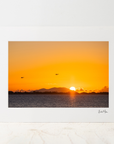 A Small photo mount featuring a vibrant orange and yellow sunset over water, displayed on a light marble surface against a neutral white wall.