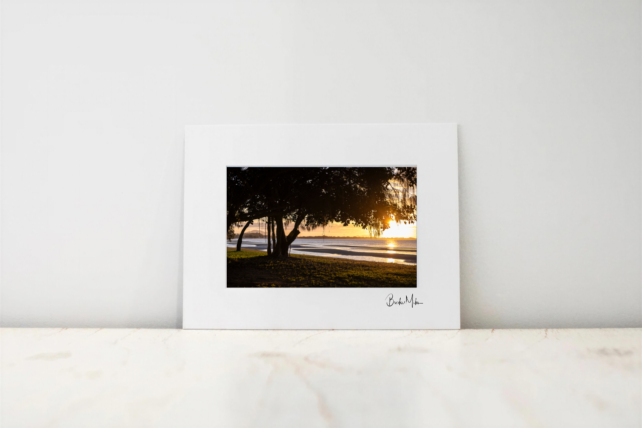A framed or matted art print leaning against a white wall on a light marble surface. The print shows a sunset seascape with dark trees on the left and golden light over the water and shoreline.