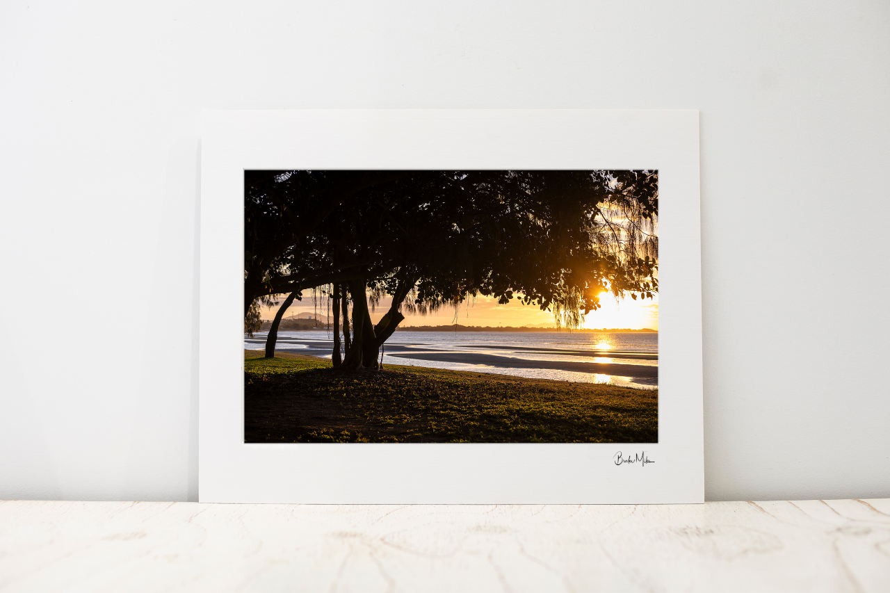 A large photo mount featuring a sunset over a shoreline with silhouetted trees, displayed against a white wall on a light marble surface.