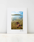 A vertical photographic art print with a wide white border, leaning against a white wall on a light marble surface. The print depicts a coastal scene with shallow blue water, distant land, and two red starfish in the foreground.
