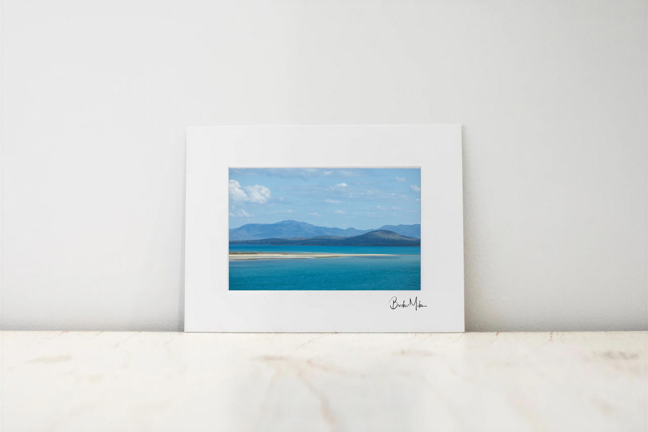 A framed landscape art print featuring a scenic view of a blue lake with mountains in the distance, displayed on a light marble surface against a white wall.