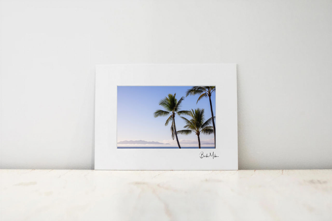 A matted art print featuring a photograph of palm trees against a blue sky, displayed on a light marble surface against a white wall.