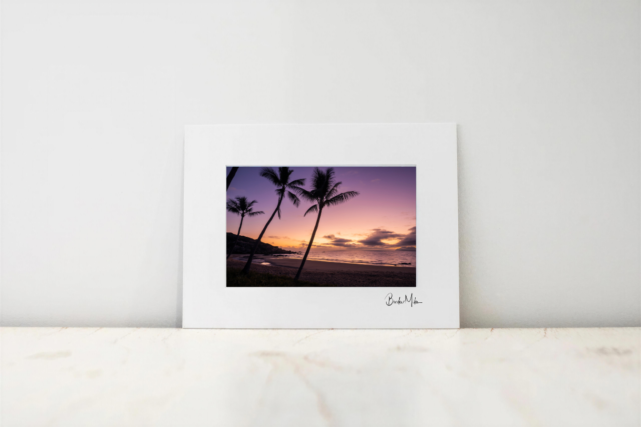 A framed art print featuring a sunset beach scene with palm tree silhouettes, displayed on a light marble surface against a white wall.