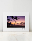 A framed art print featuring a sunset beach scene with palm tree silhouettes, displayed on a light marble surface against a white wall.