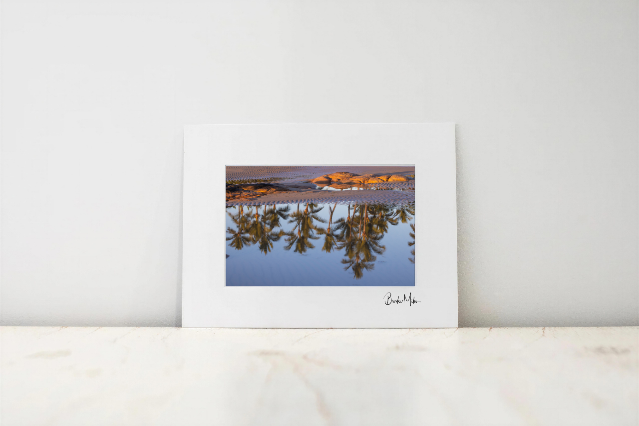 A framed art print with a wide white mat and a central landscape photograph showing a lake reflection, leaning against a white wall on a light marble surface.