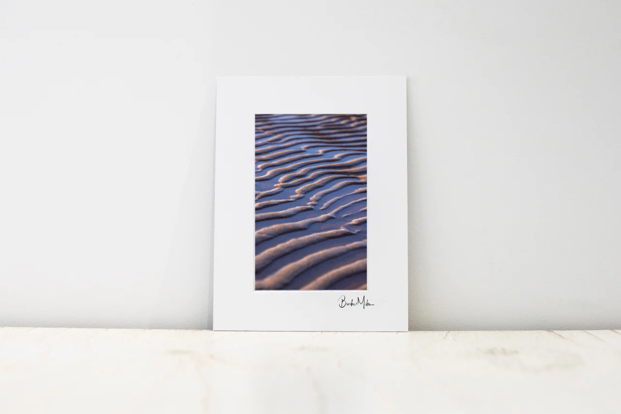 A vertical art print with a wide white border, featuring a photograph of blue and brown wavy dune patterns, leaning against a white wall on a light marble surface.