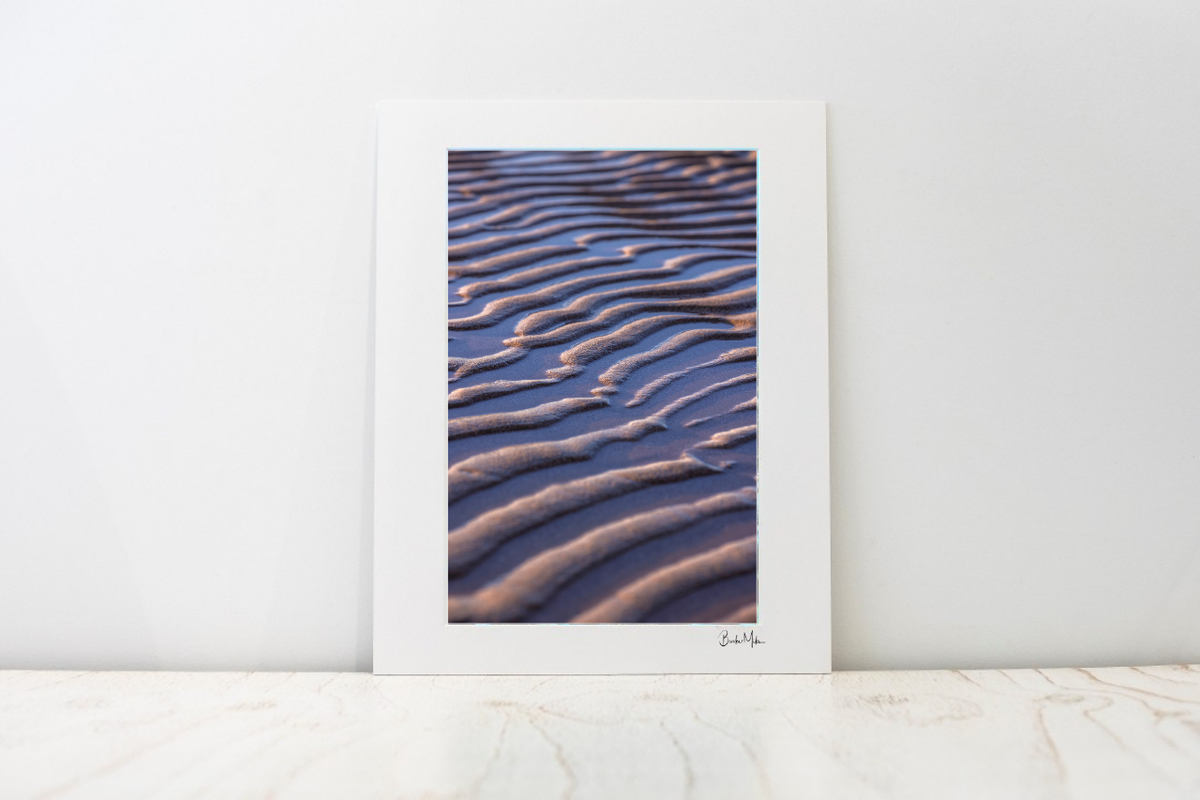 A large photo mount with a wide white border leaning against a white wall on a light marble surface. The print features a close-up photograph of rippled sand dunes in warm beige and brown tones against a deep blue-violet background.