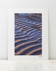 A large photo mount with a wide white border leaning against a white wall on a light marble surface. The print features a close-up photograph of rippled sand dunes in warm beige and brown tones against a deep blue-violet background.