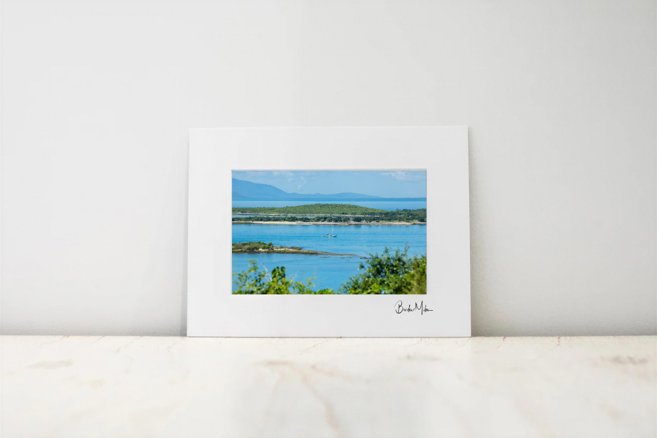 A matted art print featuring a scenic photograph of a coastal bay with blue water, green shoreline, and distant mountains, displayed on a light marble surface against a white wall.