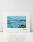 A matted art print featuring a scenic photograph of a coastal bay with blue water, green shoreline, and distant mountains, displayed on a light marble surface against a white wall.