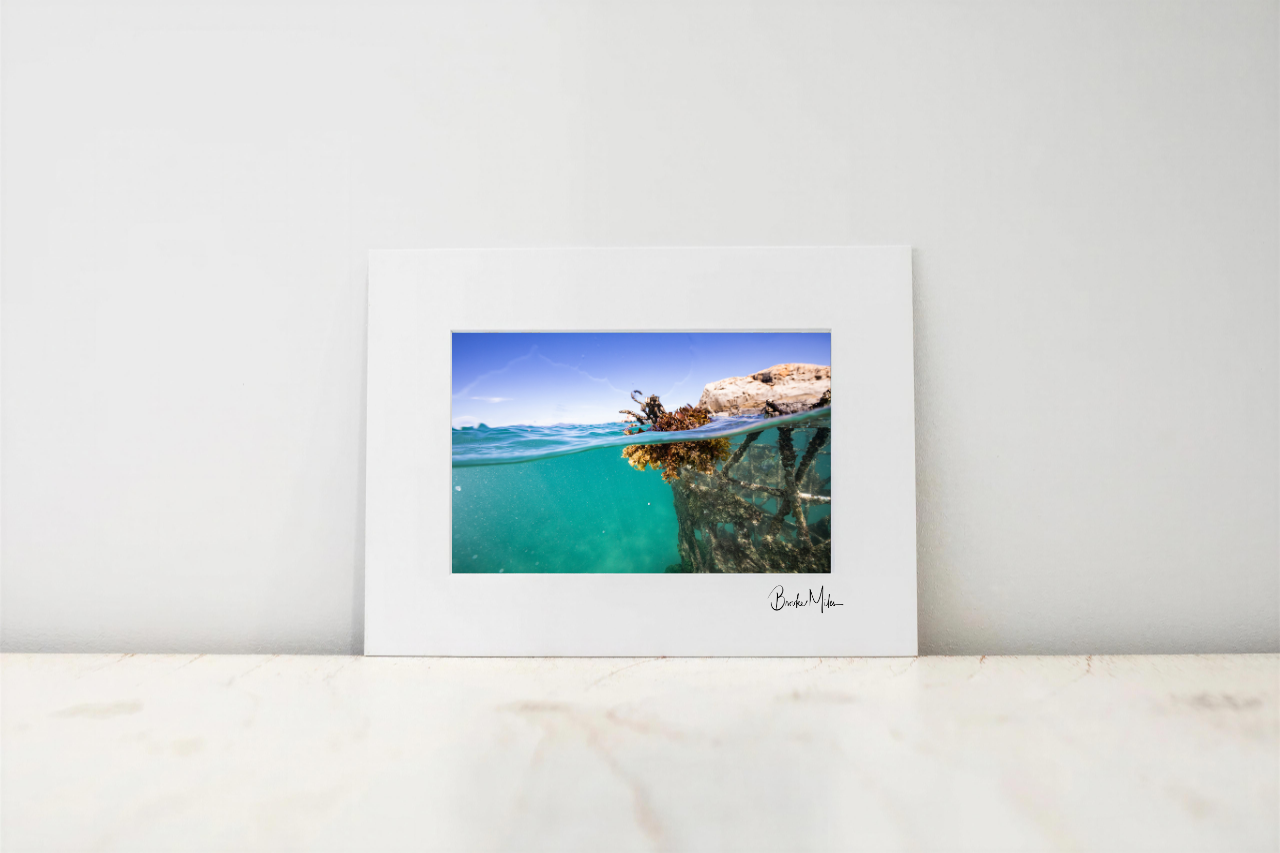 A framed photographic print leaning against a white wall on a light marble surface. The print shows a split-level ocean scene with turquoise water and a rocky shoreline under a blue sky, with a white mat and a small signature at the bottom right.