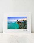 A framed photographic print leaning against a white wall on a light marble surface. The print shows a split-level ocean scene with turquoise water and a rocky shoreline under a blue sky, with a white mat and a small signature at the bottom right.