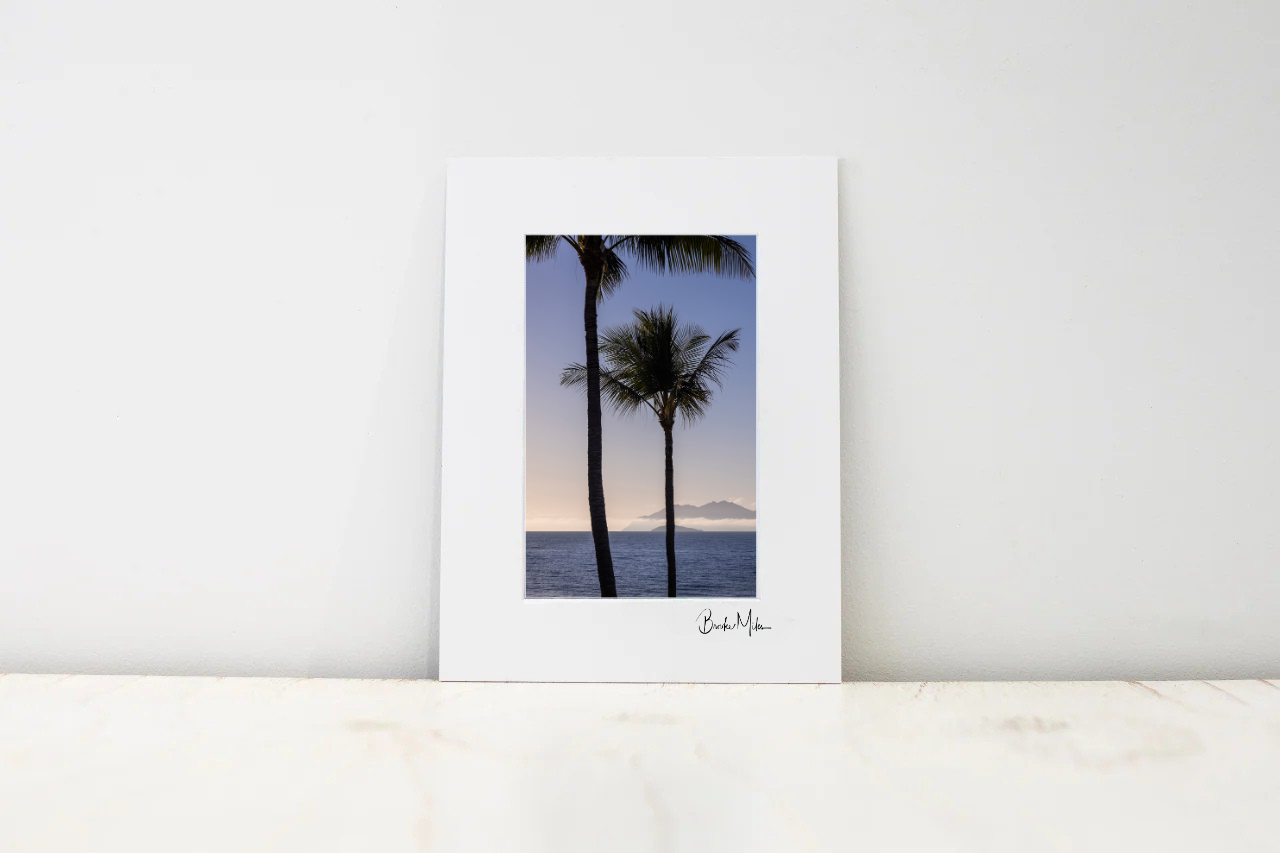 A vertical art print with a wide white border featuring a photograph of two palm trees against a sunset sky over the ocean, displayed on a light marble surface against a white wall.