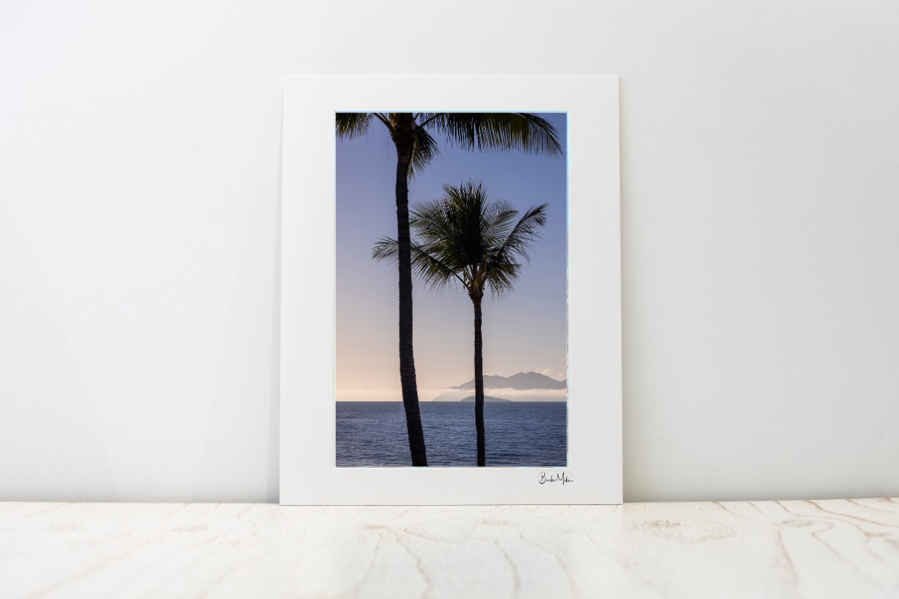 A large photo mount featuring two palm trees silhouetted against a sunset sky over the ocean, displayed on a light marble surface against a white wall.