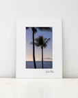 A vertical art print with a wide white border featuring a photograph of two palm trees against a sunset sky over the ocean, displayed on a light marble surface against a white wall.