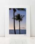 A large photo mount featuring two palm trees silhouetted against a sunset sky over the ocean, displayed on a light marble surface against a white wall.