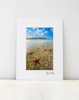 A vertical white art print with a white mat border, featuring a photograph of a beach scene with a blue sky, shallow water, and red starfish, leaning against a white wall on a light marble surface.