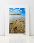 A vertical white art print with a white mat border, featuring a photograph of a beach scene with a blue sky, shallow water, and red starfish, leaning against a white wall on a light marble surface.