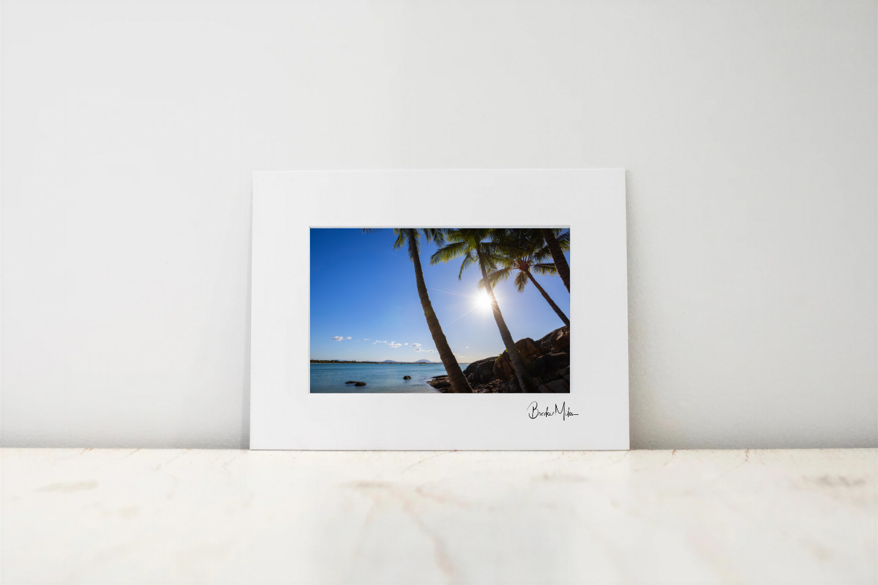 A framed or matted art print leaning against a white wall on a light marble surface. The print shows a tropical beach scene with palm trees, blue sky, water, and the sun shining, with a small signature at the bottom right.