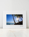 A framed or matted art print leaning against a white wall on a light marble surface. The print shows a tropical beach scene with palm trees, blue sky, water, and the sun shining, with a small signature at the bottom right.