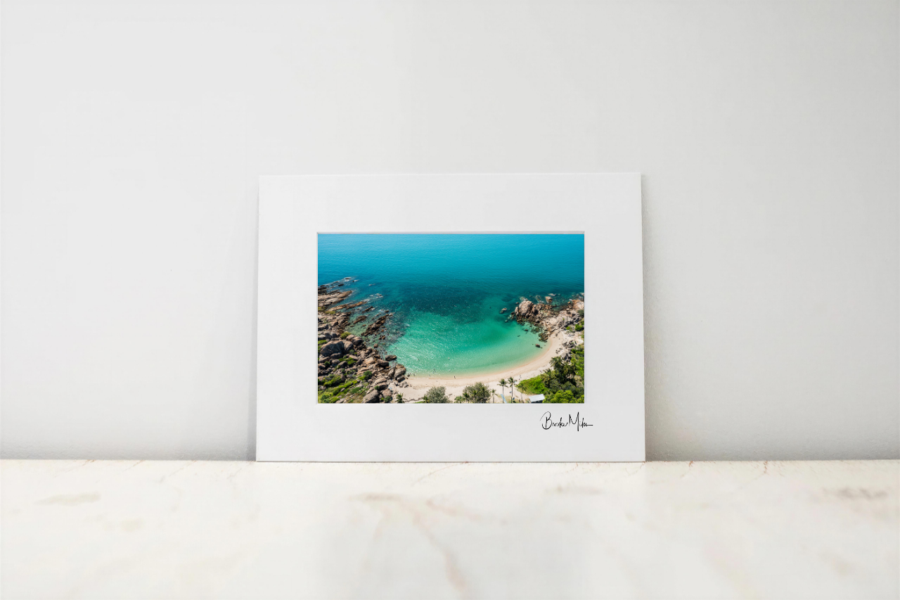 A framed photographic art print of a tropical beach cove with turquoise water, sandy shore, and rocky cliffs, displayed against a light gray wall on a white marble surface.