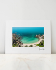 A framed photographic art print of a tropical beach cove with turquoise water, sandy shore, and rocky cliffs, displayed against a light gray wall on a white marble surface.