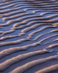 A close-up photograph of rippled wet sand with warm golden highlights and cool blue shadows, filling the frame. A small artist signature watermark appears in the bottom right corner.