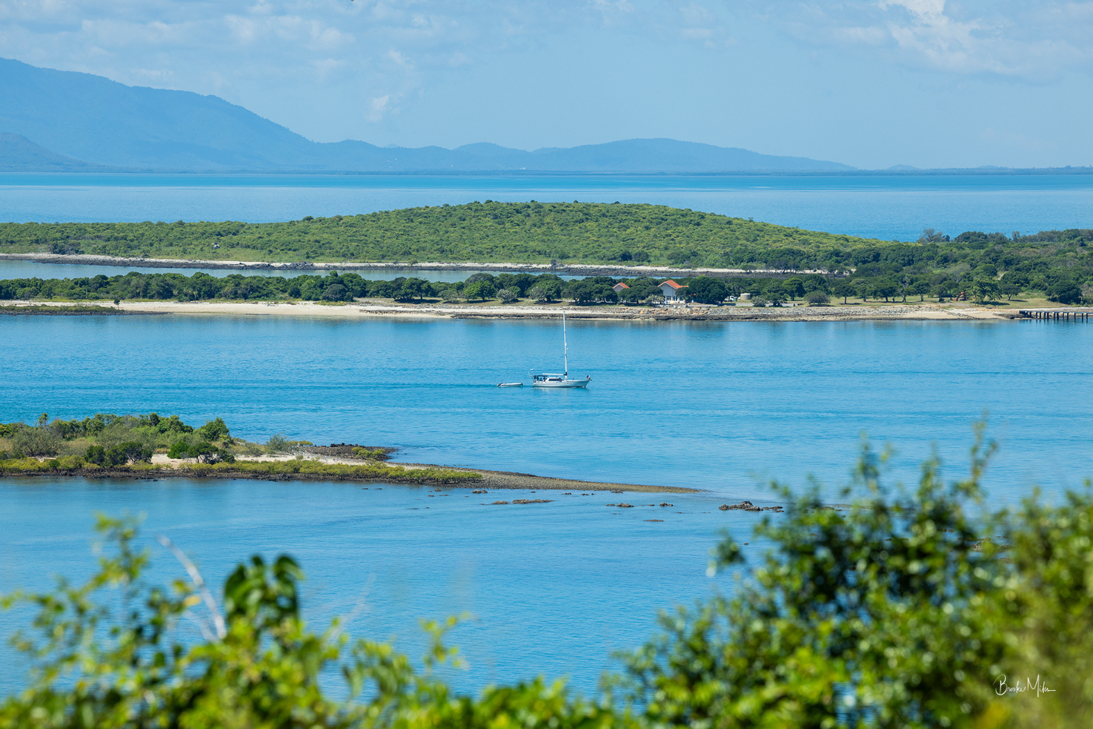 Scenic coastal view with bright blue water, a small sailboat in the distance, green islands, and mountains under a pale blue sky, framed by blurred green foliage in the foreground.