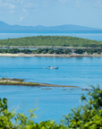 Scenic coastal view with bright blue water, a small sailboat in the distance, green islands, and mountains under a pale blue sky, framed by blurred green foliage in the foreground.