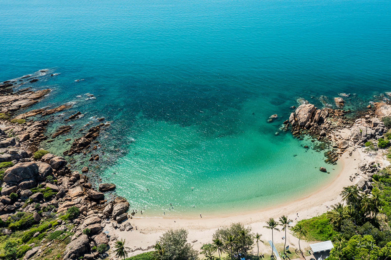 Aerial view of a tropical beach cove with turquoise and blue ocean water, rocky shoreline, sandy beach, and palm trees.