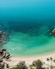 Aerial view of a tropical beach cove with turquoise and blue ocean water, rocky shoreline, sandy beach, and palm trees.