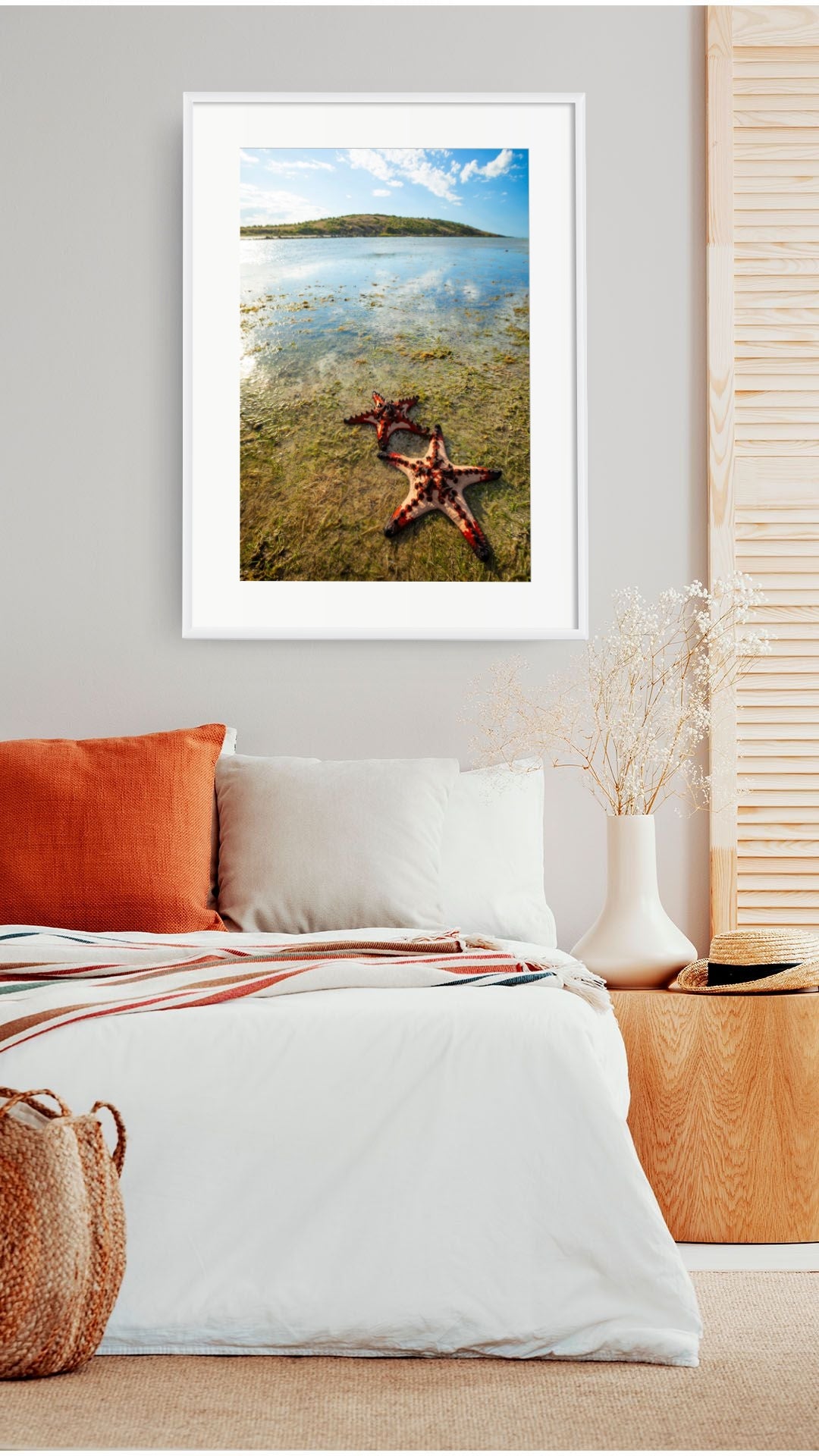 A framed wall art print showing a starfish on a sandy shore with shallow blue water and distant land, displayed above a bed in a styled bedroom interior.