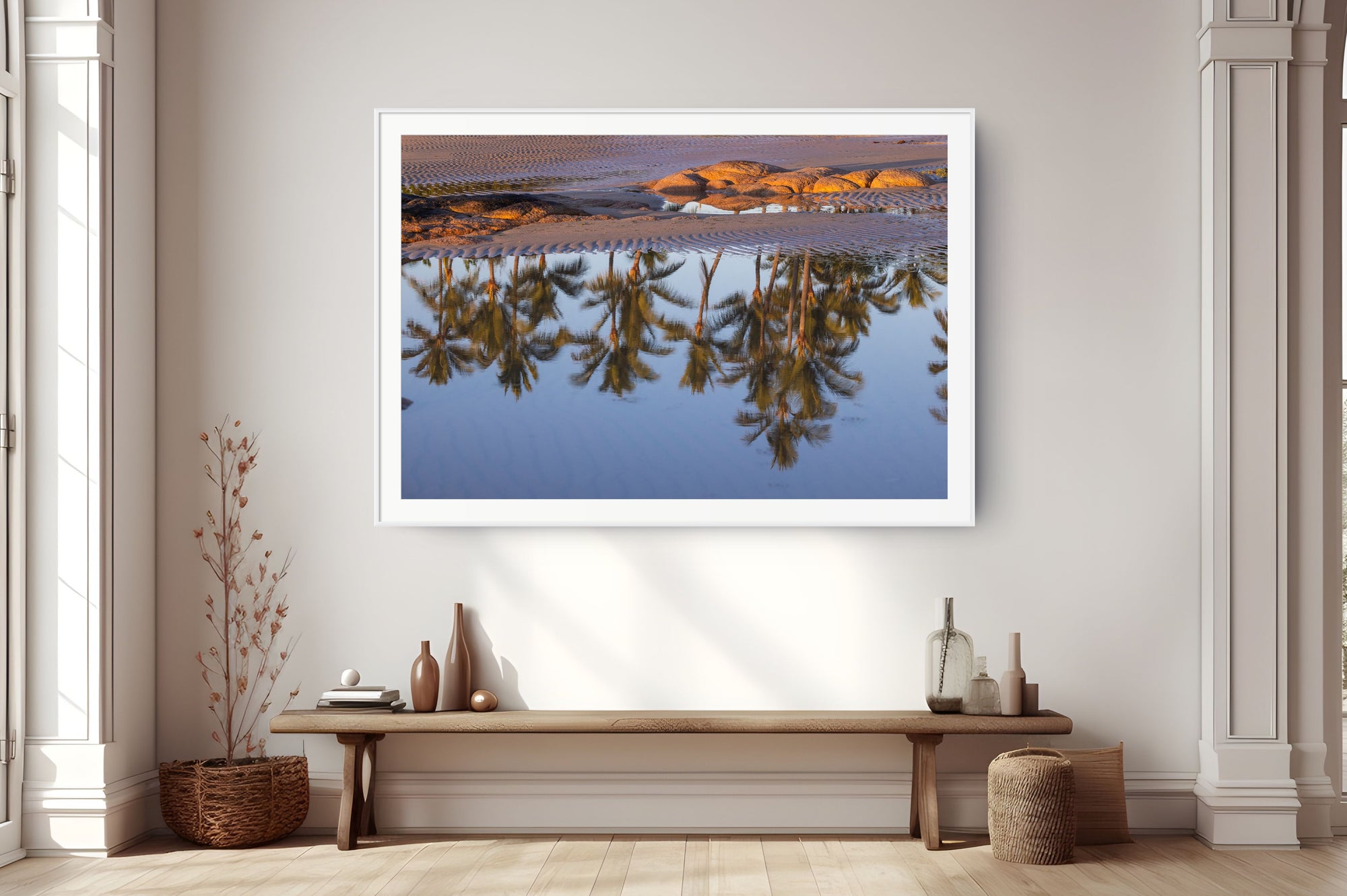 A framed landscape art print depicting desert dunes reflected in calm water with palm trees, displayed on a wall above a wooden bench in a modern, sunlit room.