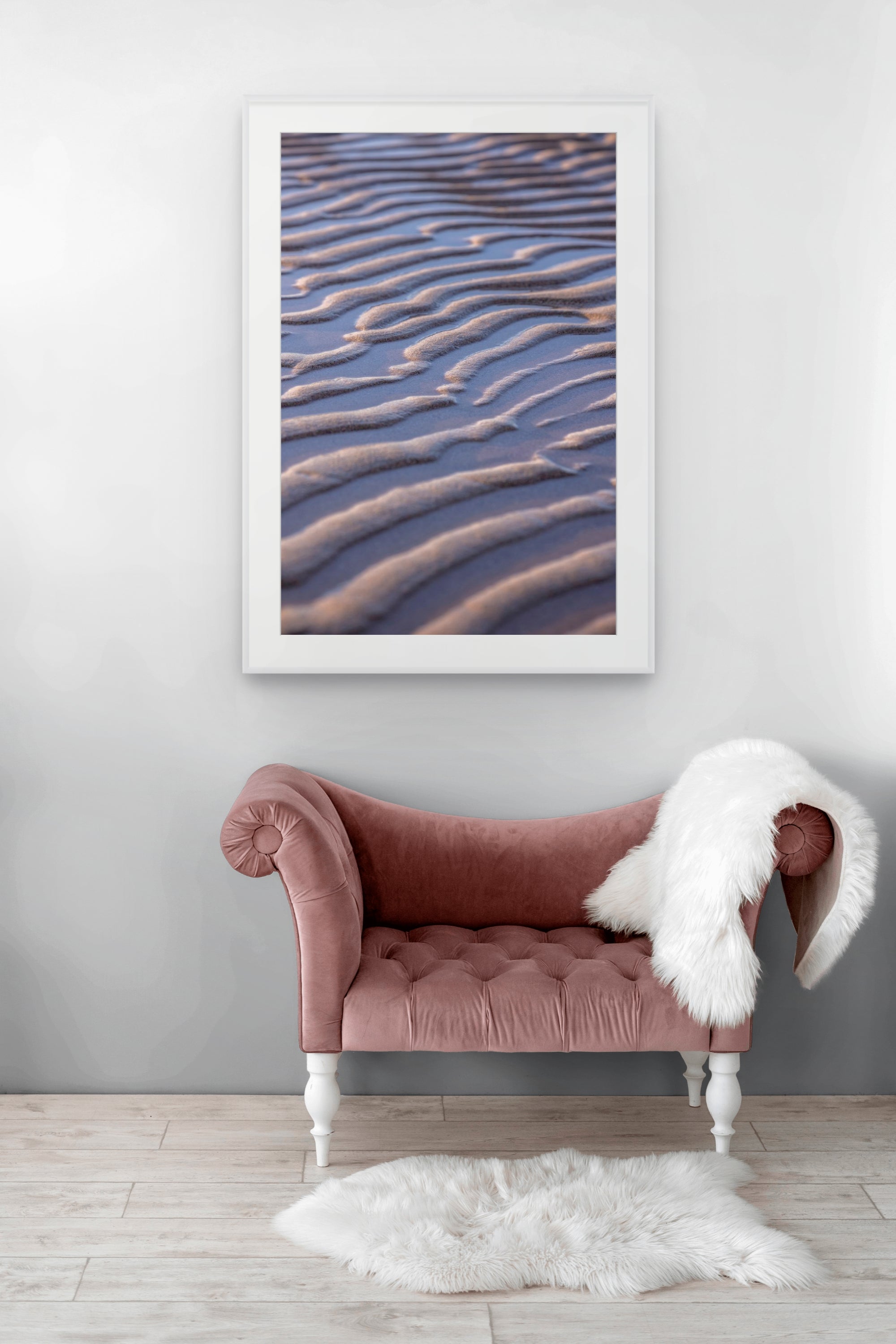 A framed wall art print showing a close-up of rippled sand dunes in warm beige and dusty blue tones, displayed above a dusty rose velvet chaise lounge with a white faux-fur throw on a light wood floor.
