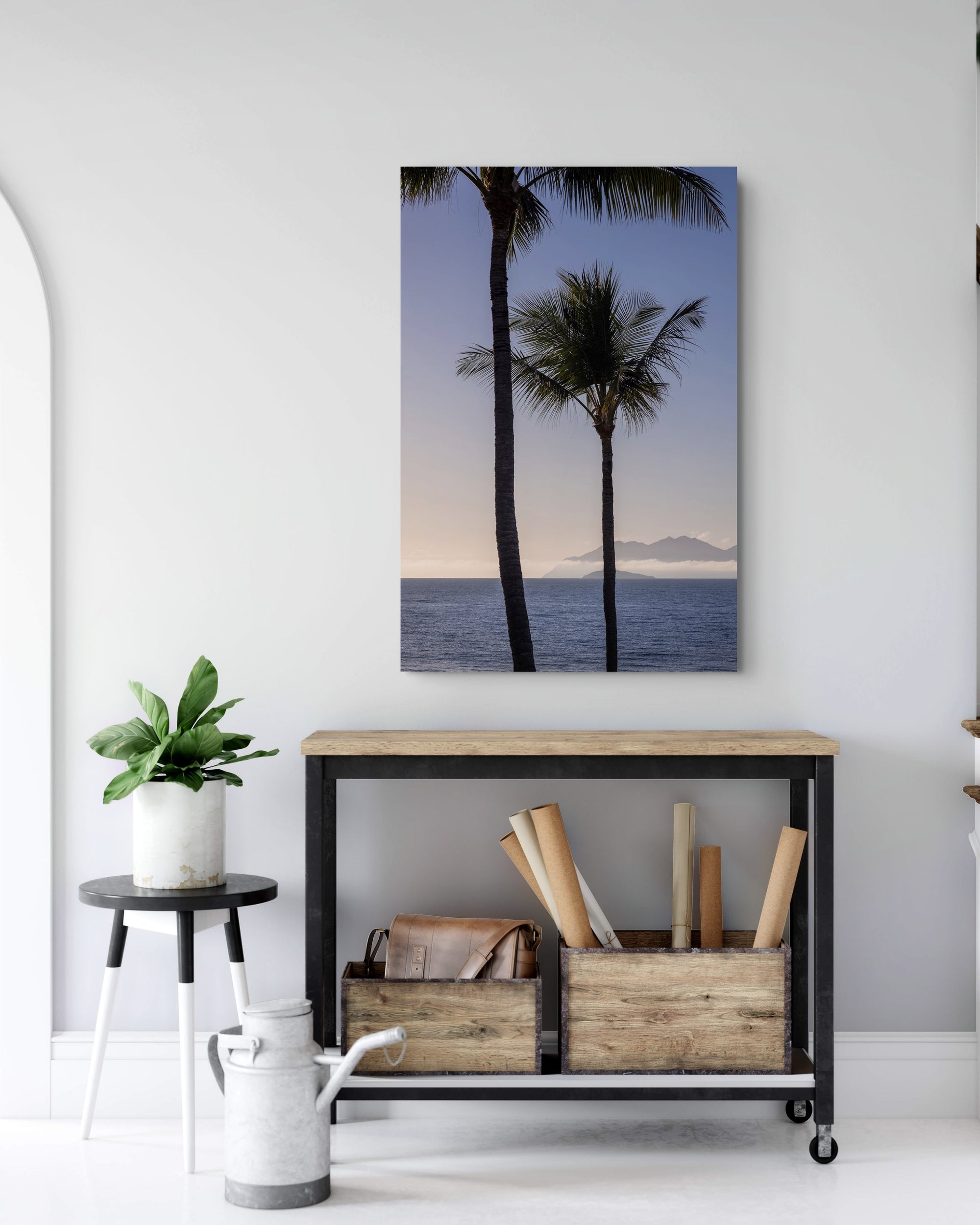 A vertical canvas wall art print depicting two palm trees over a calm ocean at sunset, styled in a modern room above a black metal console table.