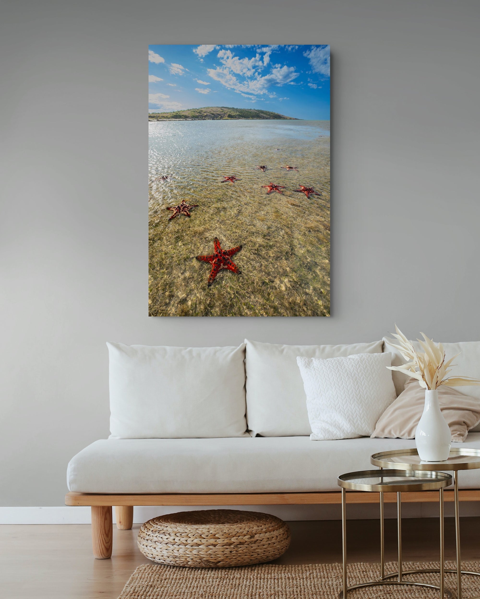 A vertical canvas wall art print depicting a beach scene with red starfish in shallow water, displayed above a white sofa in a modern living room setting.