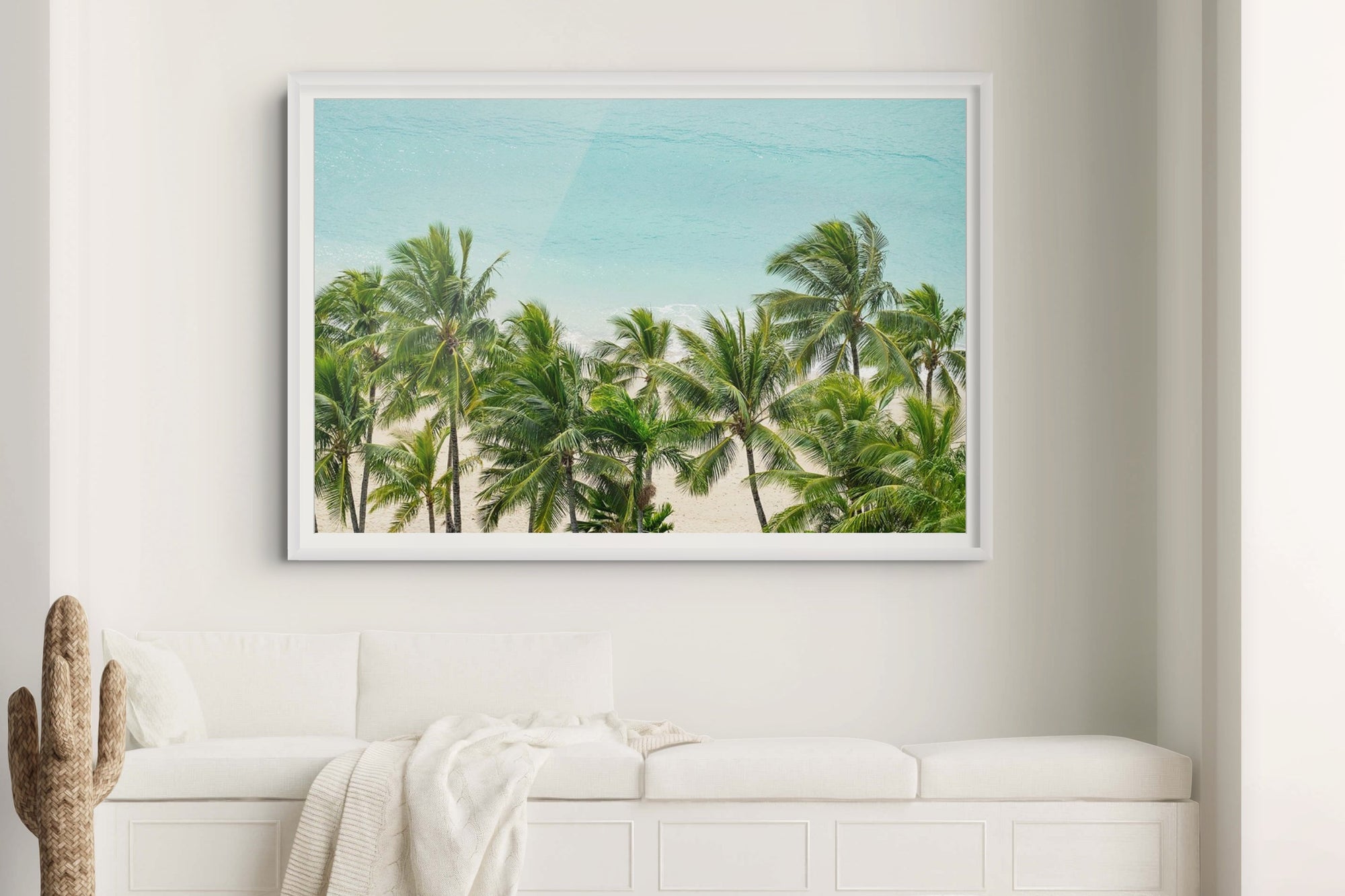 Framed tropical beach scene with palm trees on a white wall above a bench.