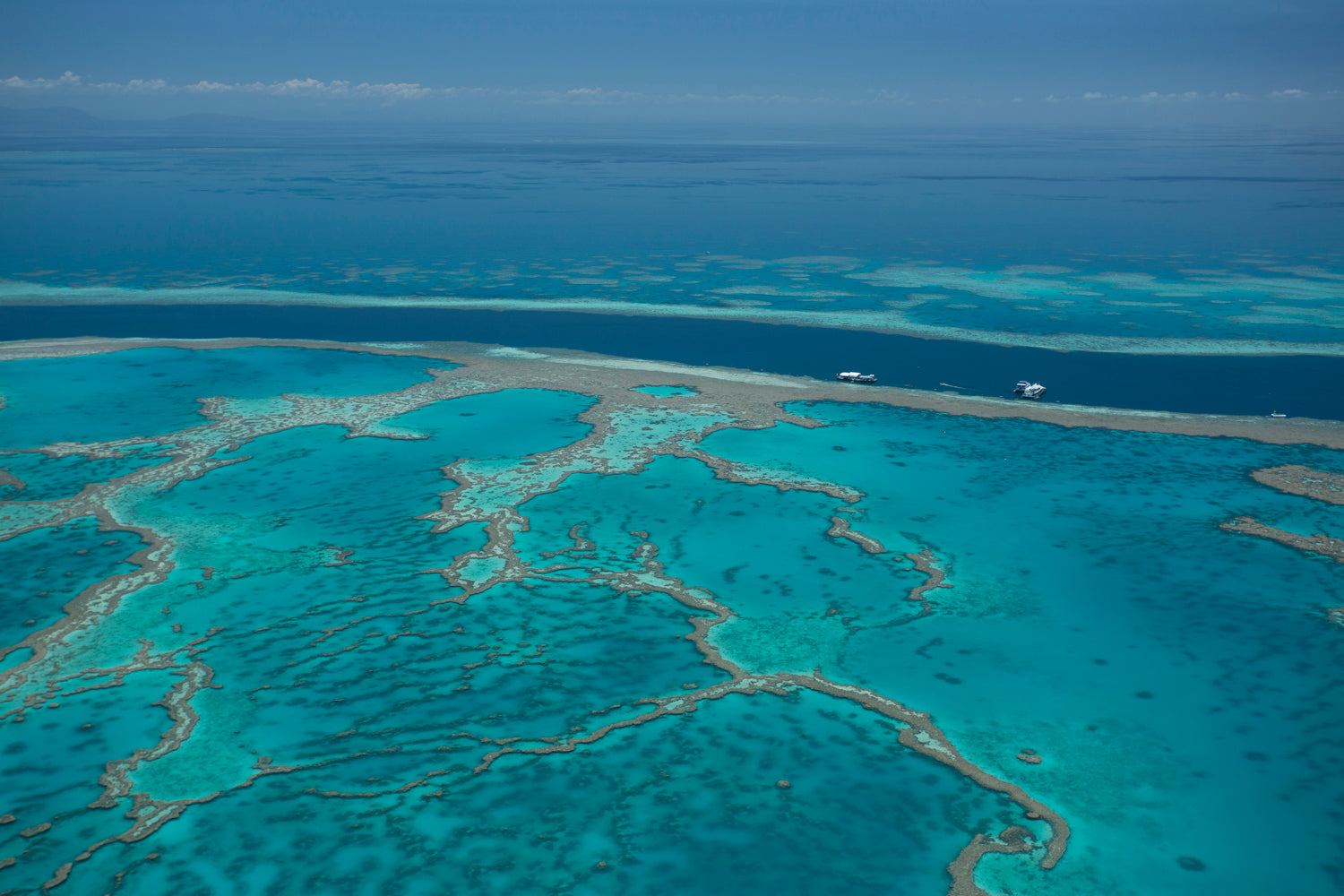 aerial of veiny coral reef amongst turquiose water deep water beyond with large boats on anchor