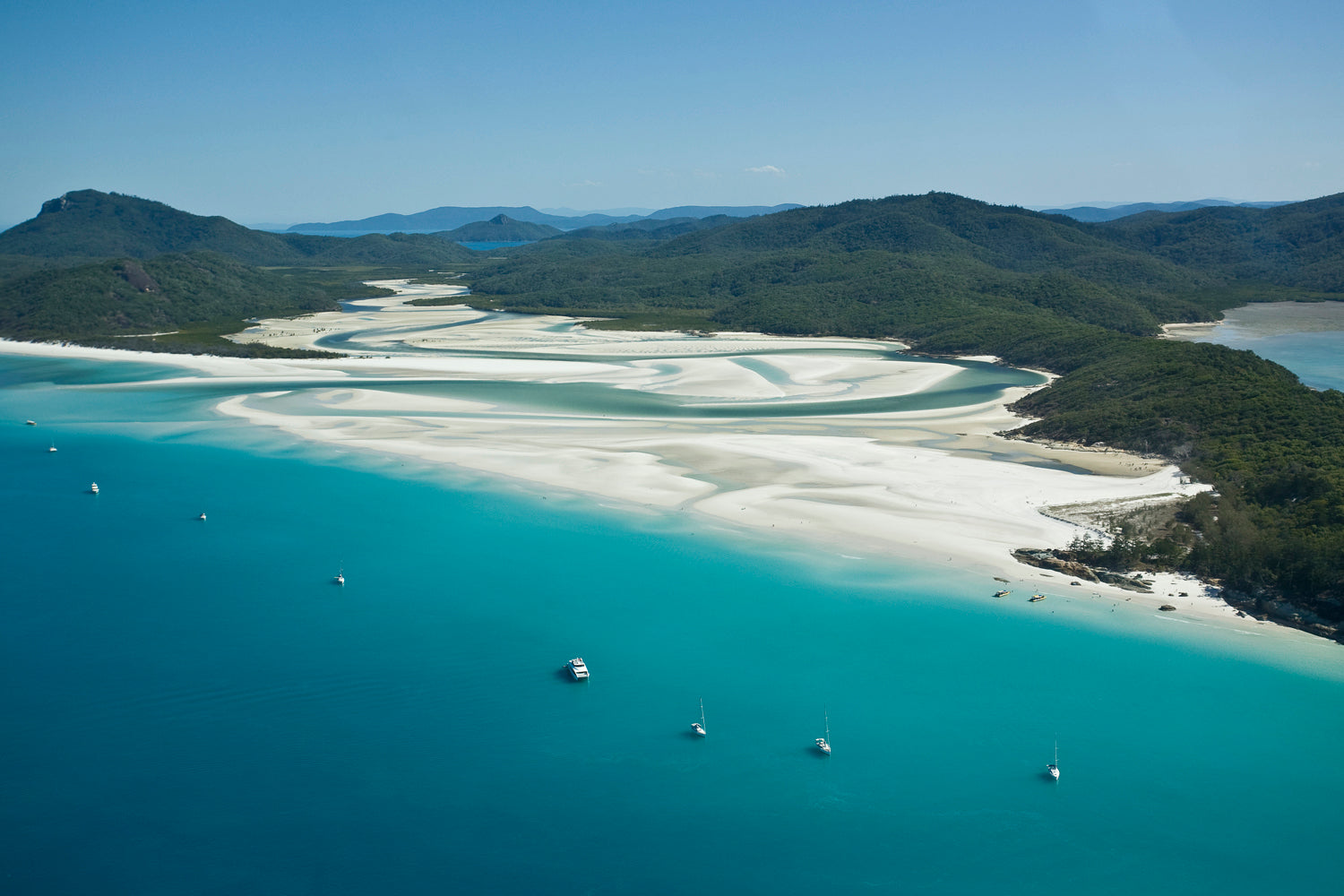 neat row of anchored vessels along a white sandy beach with a swirling turquiose water river running out the ocean
