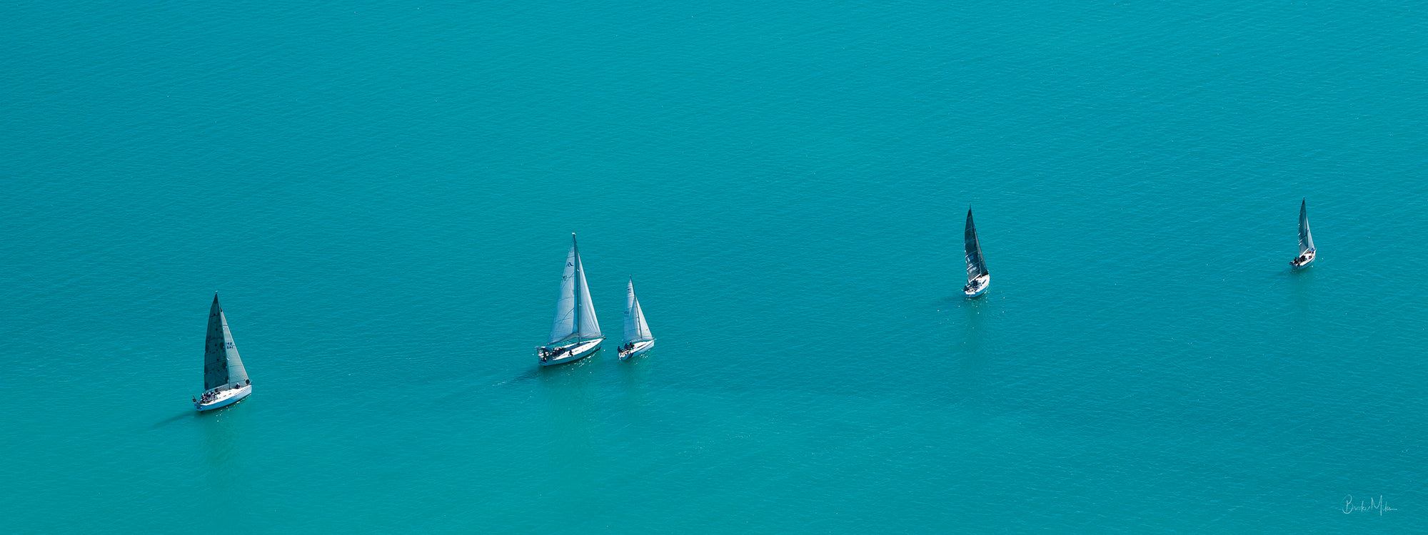 5 sailing vessels with their sails raised in a line, one behind the other with blue ocean surrounding them
